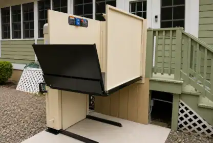A beige wheelchair lift is installed next to green steps, providing access from the ground level to a porch door of a house.