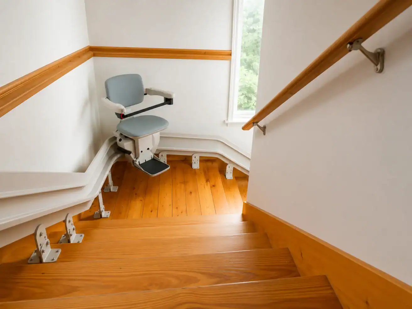 A stairlift is installed along a wooden staircase with a curved rail, next to a handrail and a window providing natural light.