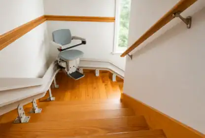 A stairlift is installed along a wooden staircase with a curved rail, next to a handrail and a window providing natural light.