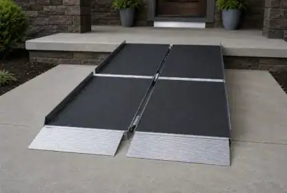 A portable, foldable wheelchair ramp is set up on a concrete entrance, leading to a raised doorway with two potted plants by the door.