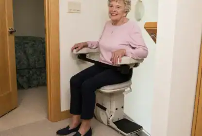 An older woman sits on a stair lift chair at the bottom of a staircase in a home, smiling at the camera.