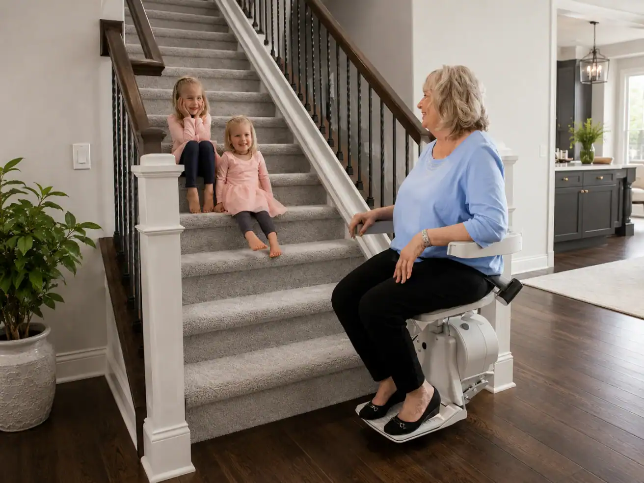 Older woman sits on a stair lift at the bottom of carpeted stairs, smiling at two young girls sitting on the steps in a modern, bright home.