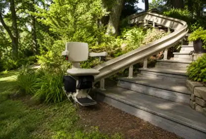 Outdoor stairlift installed along a curved wooden staircase surrounded by green plants and trees in a garden setting.