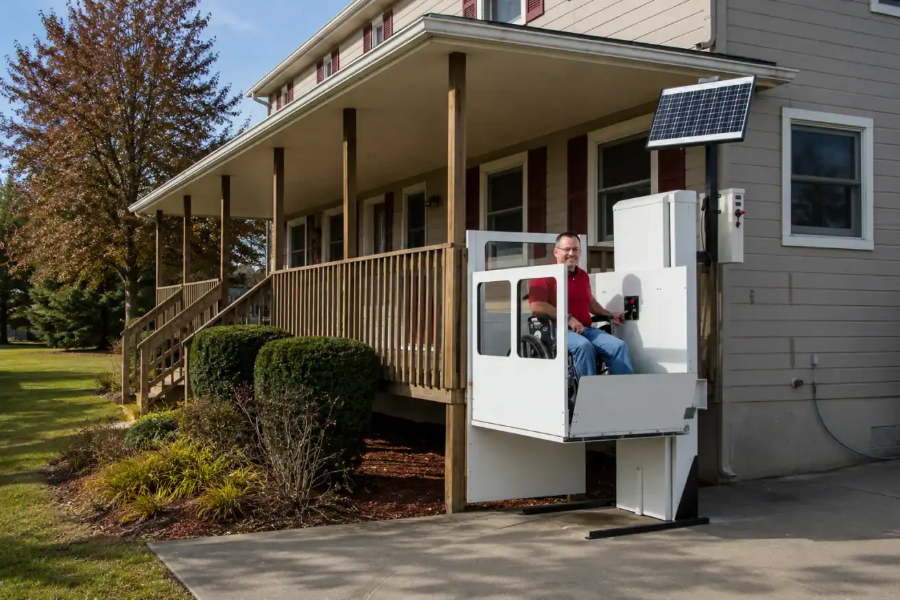 A man uses an outdoor wheelchair lift with solar panel at the entrance of a house with a porch and ramp.