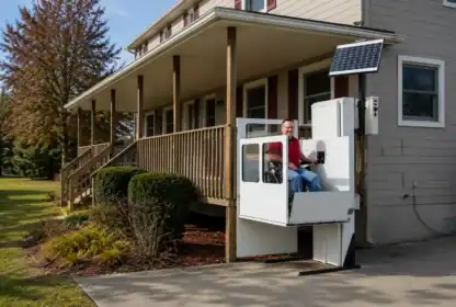 A man uses an outdoor wheelchair lift with solar panel at the entrance of a house with a porch and ramp.