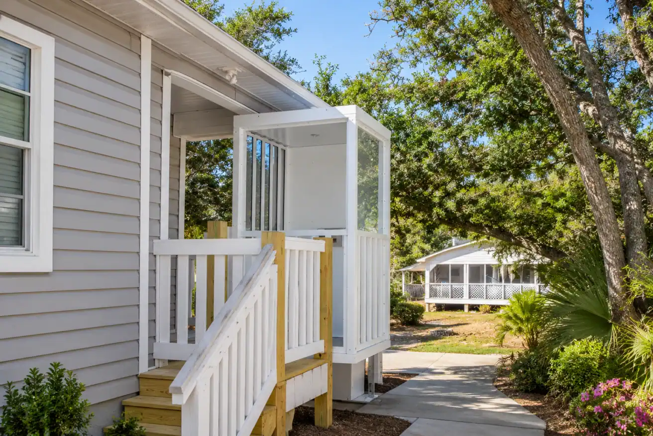 A residential wheelchair lift is installed next to a small porch with stairs, surrounded by trees and a walkway.