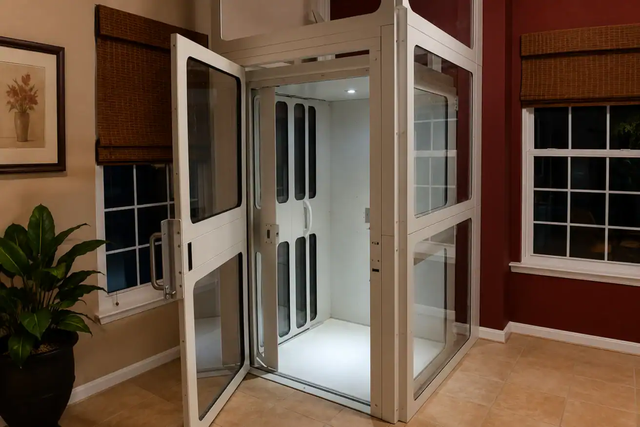 A residential elevator with glass doors stands open in a room with tiled floors, red and beige walls, windows with brown blinds, and a potted plant on the left.