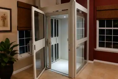 A residential elevator with glass doors stands open in a room with tiled floors, red and beige walls, windows with brown blinds, and a potted plant on the left.