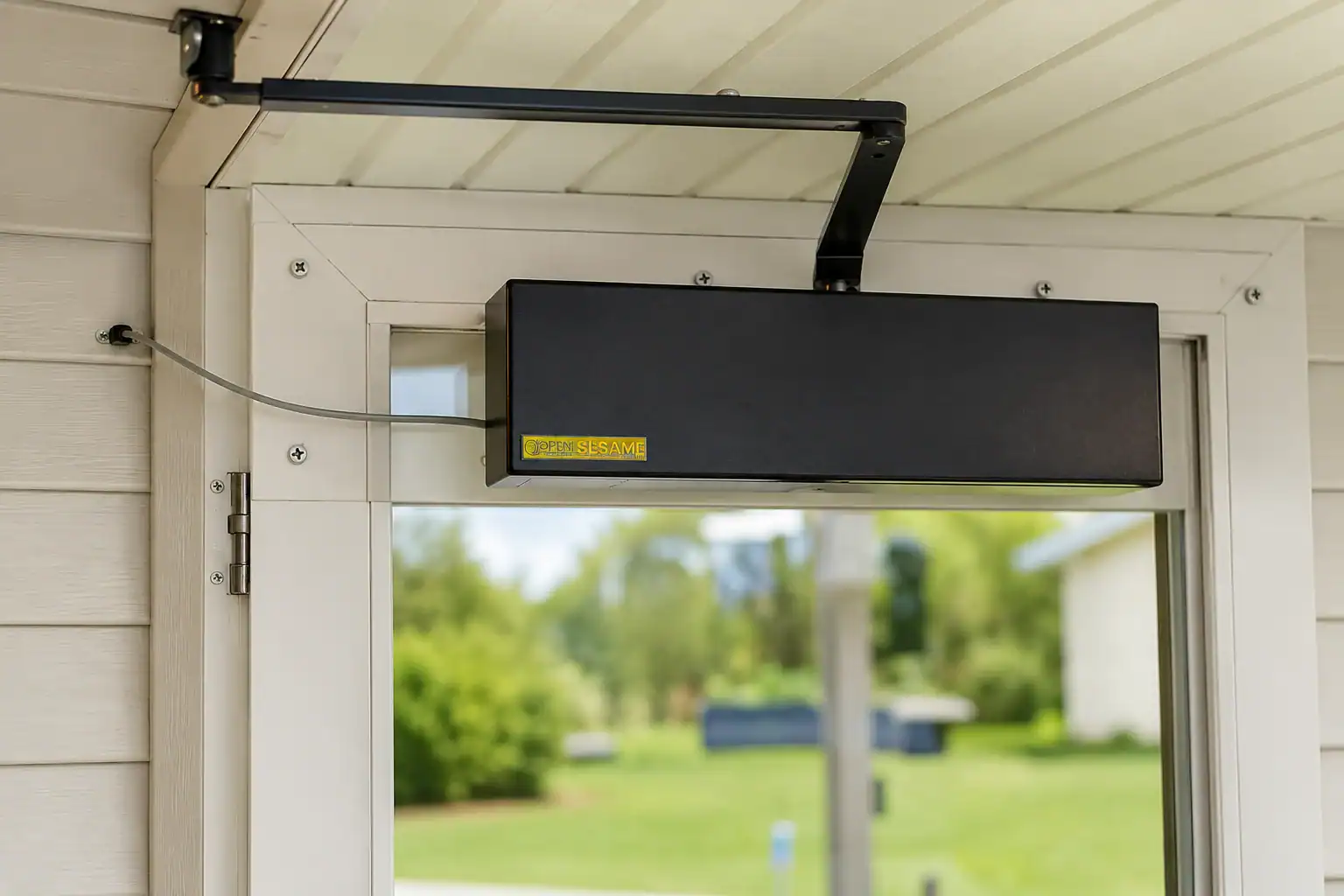 A black door operator with a yellow "Open Sesame" label is mounted above a glass door on a white porch ceiling, wired to the door frame.