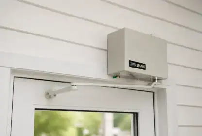 A gray automatic door opener labeled "Open Sesame" is mounted above a white framed glass door on a paneled wall.