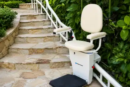 Outdoor stairlift with a padded seat and armrests installed beside a stone staircase, surrounded by green plants and bushes.