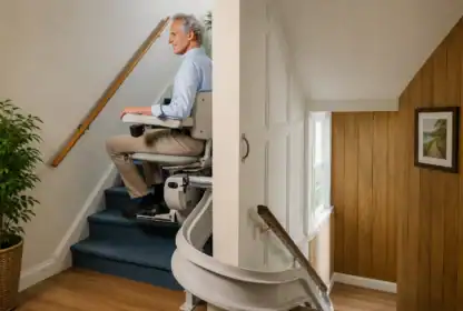 An older man rides a stairlift up a carpeted staircase in a home. The stairlift curves at the bottom, and the walls are decorated with plants, wood paneling, and framed pictures.