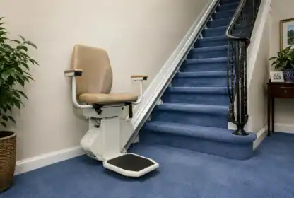 A stairlift with a beige seat is installed beside a blue carpeted staircase. A green plant and a wooden table are visible nearby against cream-colored walls.