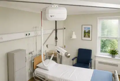 Hospital room with a patient bed, blue chair, window, medical lift system overhead, medical equipment, and a potted plant on the windowsill.