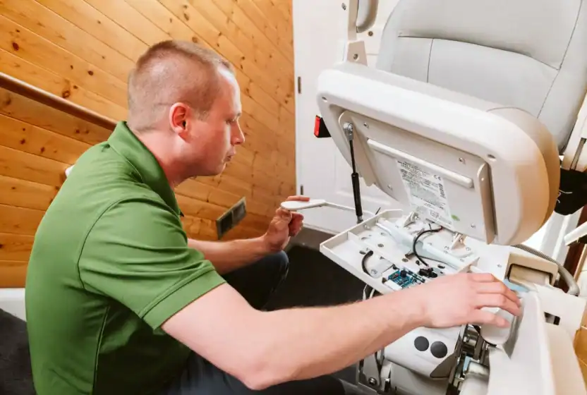 A technician in a green shirt repairs a stair lift, examining its open control panel and wiring on a wooden wall staircase.