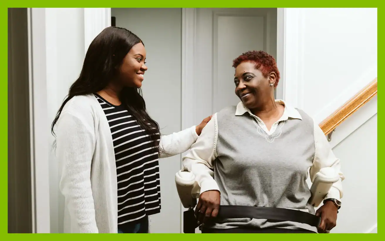 A younger woman stands beside an older woman seated on a stair lift, both smiling at each other in a hallway with stairs visible in the background.