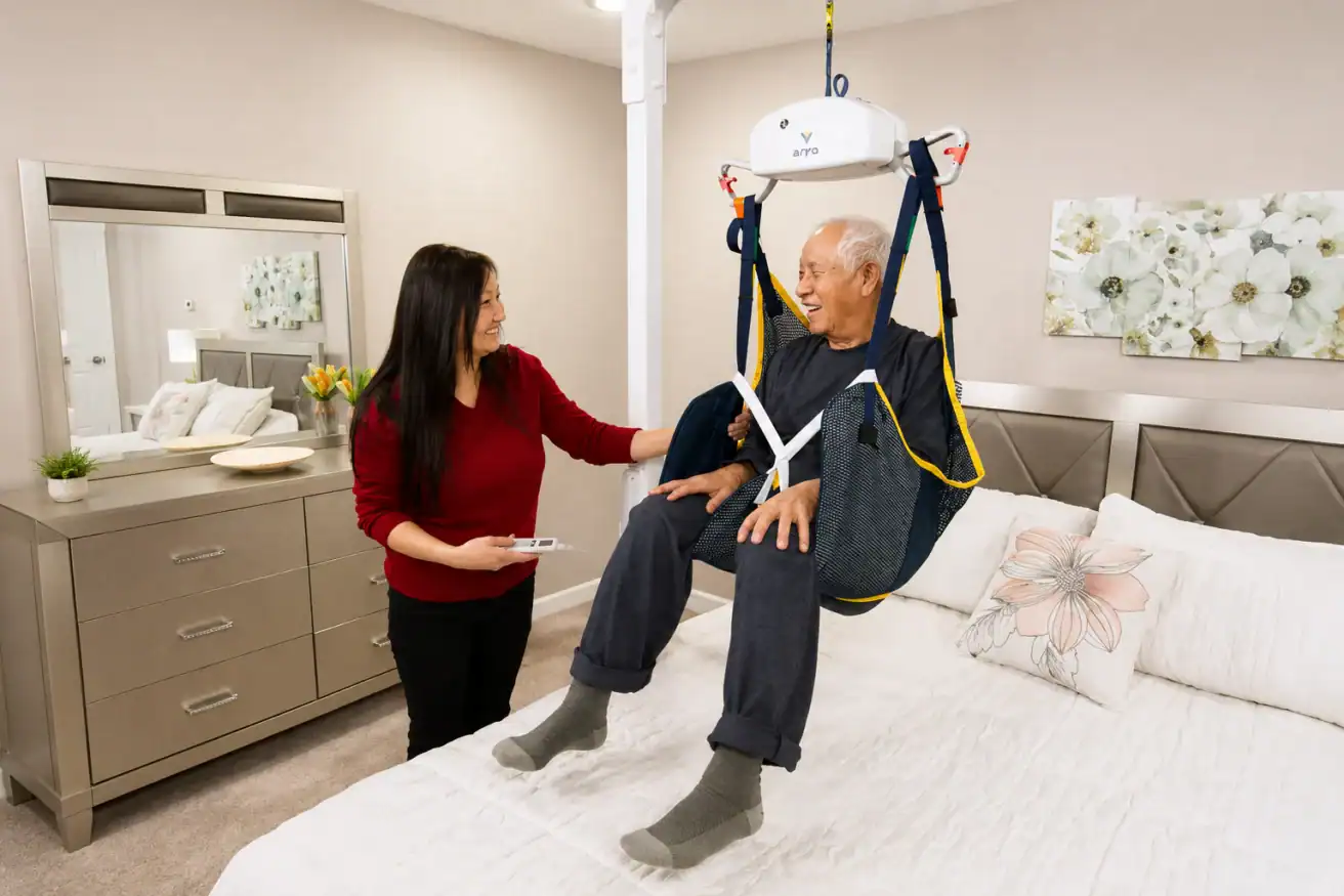 A woman assists an elderly man using a ceiling lift system over a bed in a bedroom. Both appear calm and are engaged in conversation.