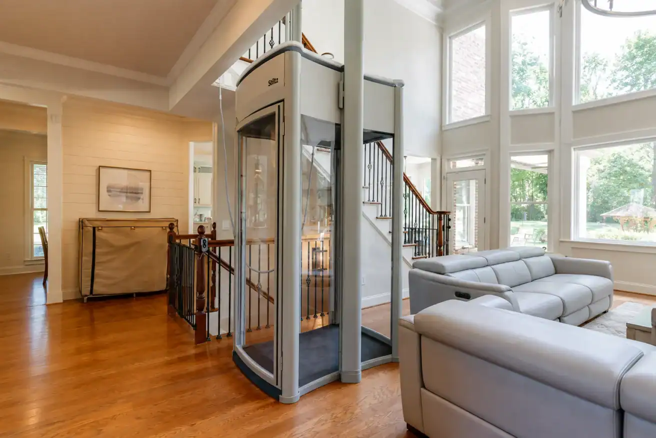 A small glass elevator is installed in a bright living room with hardwood floors, large windows, and a gray sectional sofa.