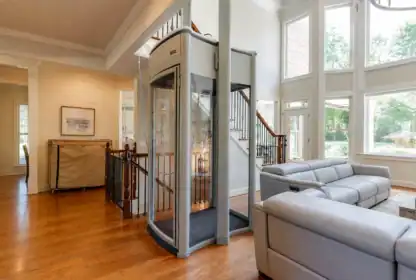 A small glass elevator is installed in a bright living room with hardwood floors, large windows, and a gray sectional sofa.