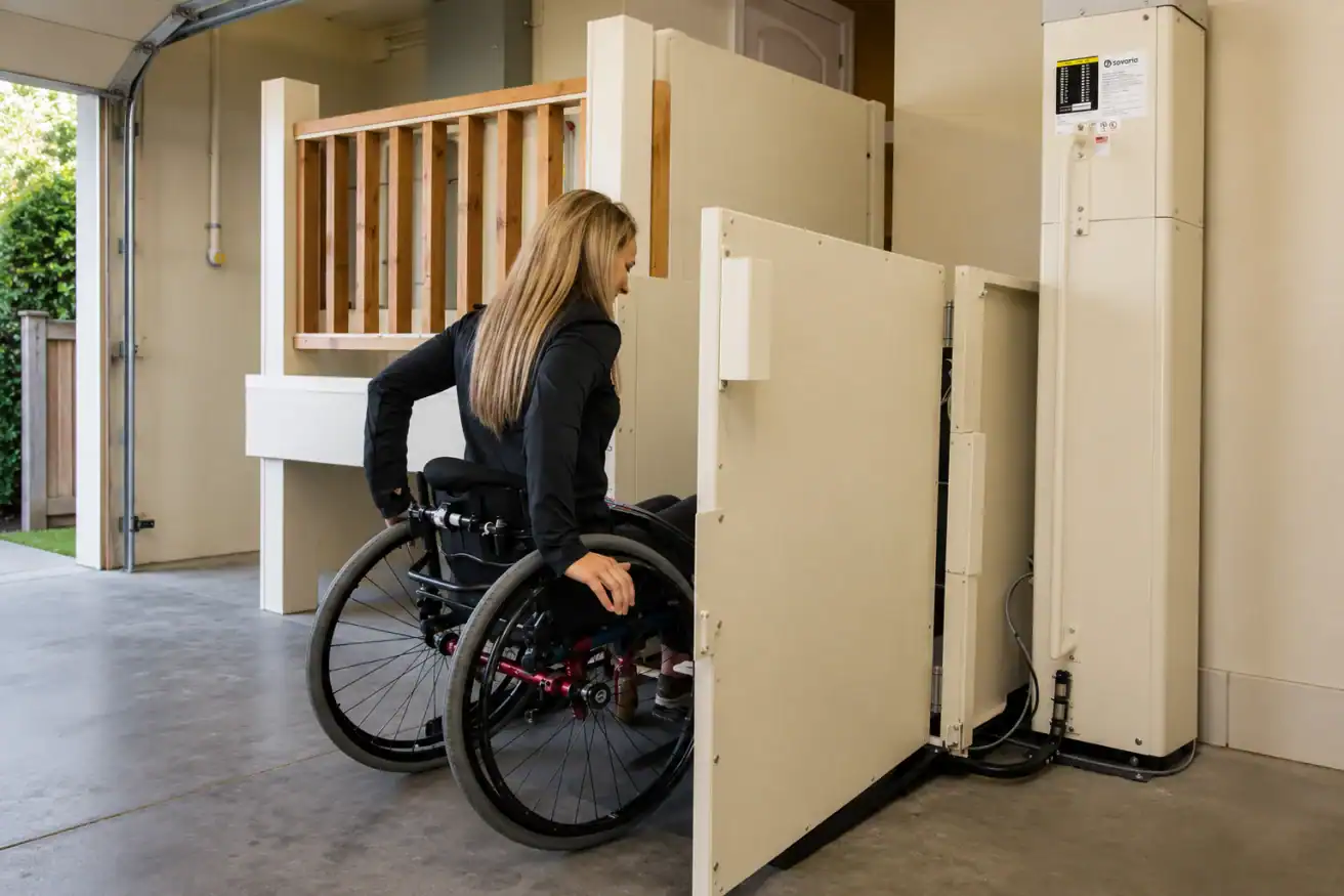 A woman in a wheelchair uses a vertical platform lift to access an elevated area inside a residential garage.