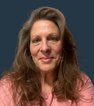 A woman with long brown hair wearing a pink top is smiling slightly, looking at the camera, with a plain dark blue background.