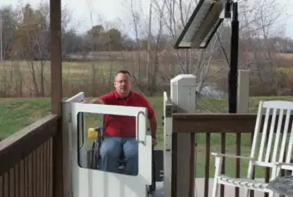 A man in a wheelchair uses a vertical platform lift to access a porch with a rocking chair. Trees and a field are visible in the background.