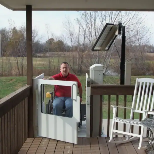A man in a wheelchair uses a vertical platform lift to access a porch with a rocking chair. Trees and a field are visible in the background.