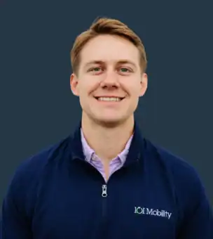 A person smiling at the camera, wearing a navy quarter-zip jacket with an 101 Mobility logo, against a plain dark background.