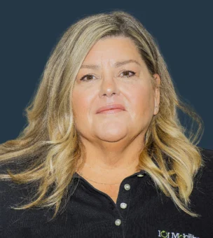 A woman with long blonde hair wearing a black collared shirt stands in front of a plain dark background, looking directly at the camera.