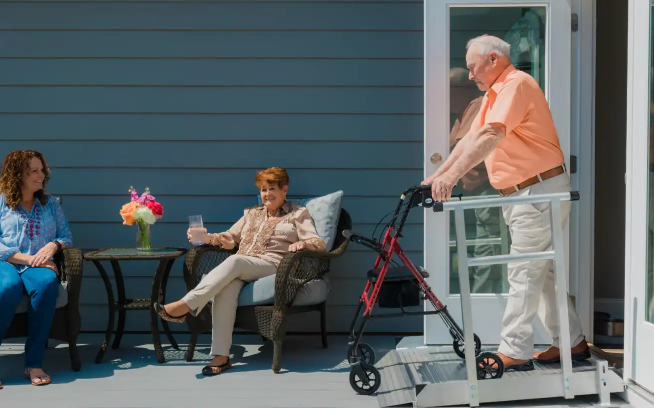 An older man uses a walker to exit a house onto a porch, where two women sit and talk at a small table with a flower vase and drinks.
