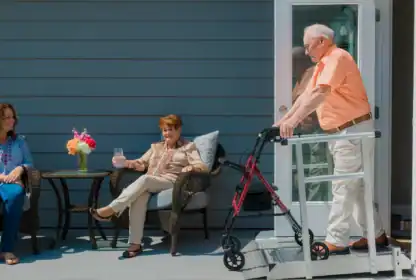 An older man uses a walker to exit a house onto a porch, where two women sit and talk at a small table with a flower vase and drinks.