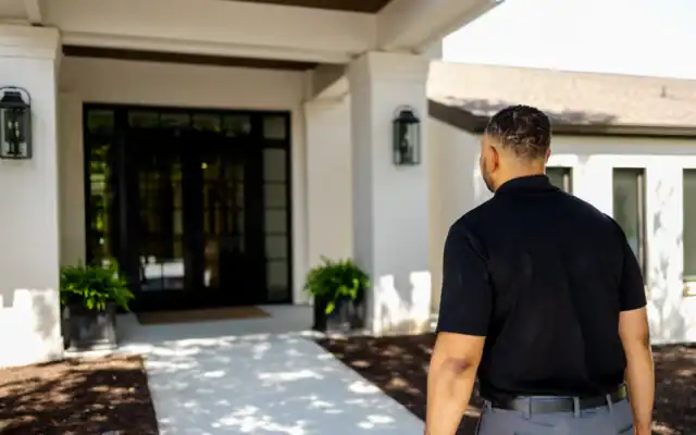 A man with short hair, wearing a black shirt and gray pants, walks toward the entrance of a modern house with glass doors and potted plants.