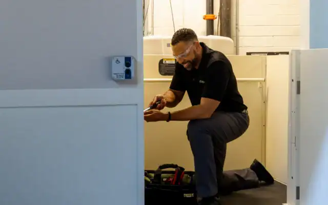 A man wearing safety glasses kneels on one knee while working with tools inside a utility area with a control panel visible on the wall.