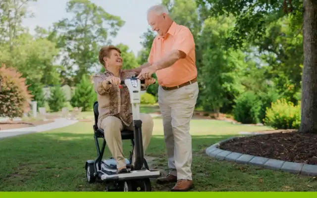 An older woman sits on a mobility scooter while an older man stands beside her, both smiling, in a sunny park with trees and grass.