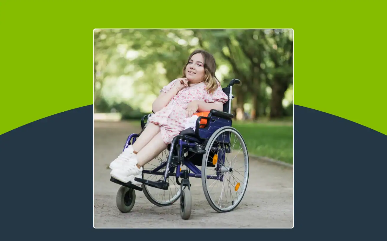 A young woman in a pink dress sits in a wheelchair outdoors on a paved path, surrounded by green trees and sunlight.