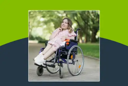 A young woman in a pink dress sits in a wheelchair outdoors on a paved path, surrounded by green trees and sunlight.