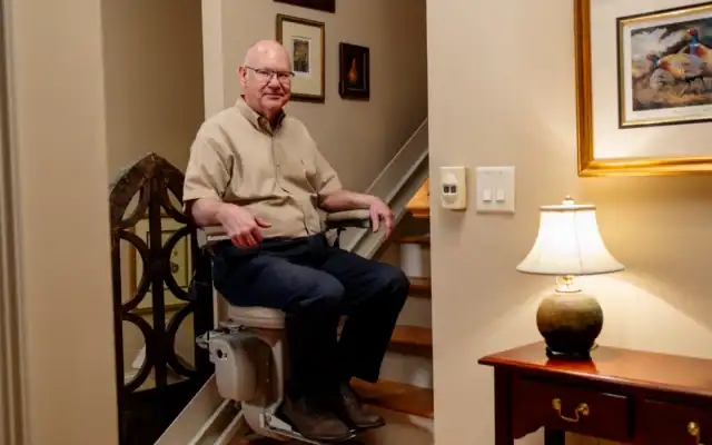 Older man sits on a stair lift at the base of a staircase in a warmly lit home, surrounded by framed art and a lamp on a side table.
