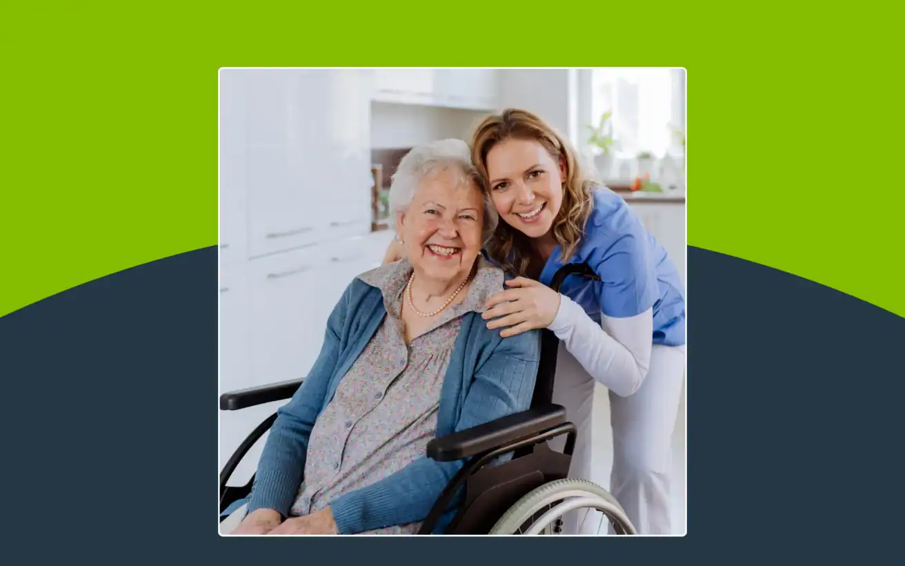 Smiling elderly woman in a wheelchair and a young caregiver in scrubs pose together in a kitchen, both looking at the camera.