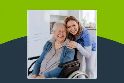 Smiling elderly woman in a wheelchair and a young caregiver in scrubs pose together in a kitchen, both looking at the camera.