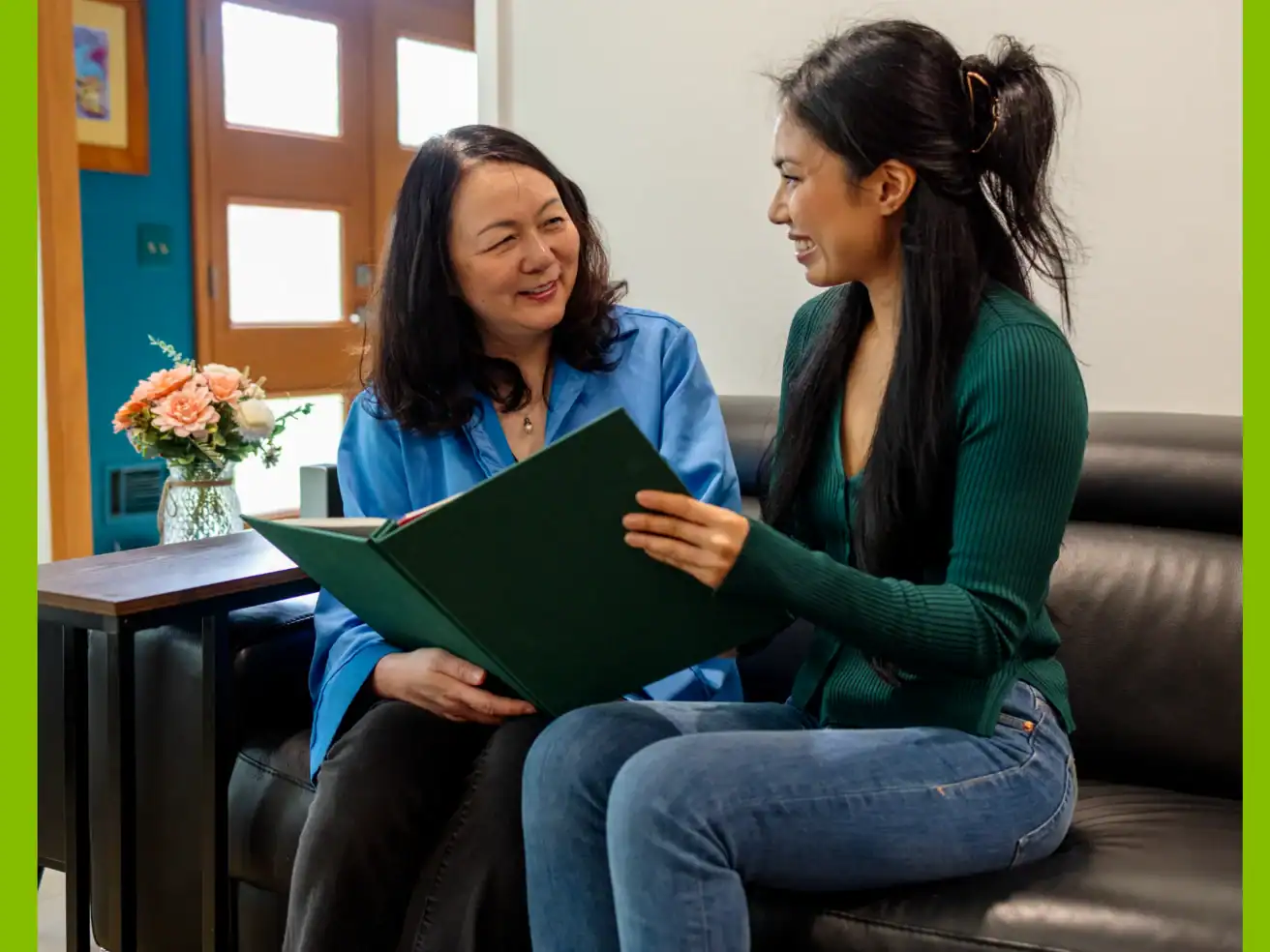 Two women sit on a couch, smiling and looking at a large green folder together. A vase of flowers sits on the table next to them.