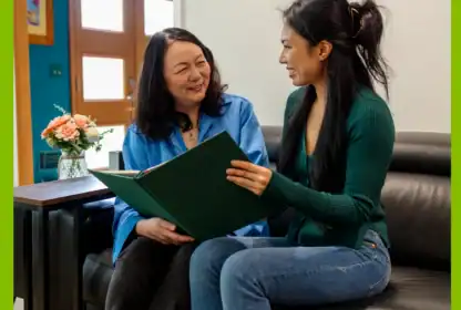 Two women sit on a couch, smiling and looking at a large green folder together. A vase of flowers sits on the table next to them.