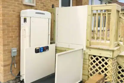 A residential wheelchair lift is installed next to a wooden deck and brick house, providing accessible entry from the yard to the porch.
