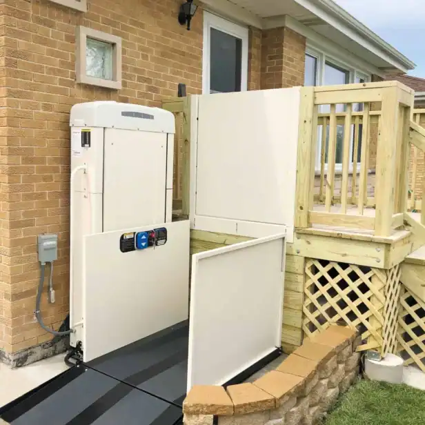 A residential wheelchair lift is installed next to a wooden deck and brick house, providing accessible entry from the yard to the porch.