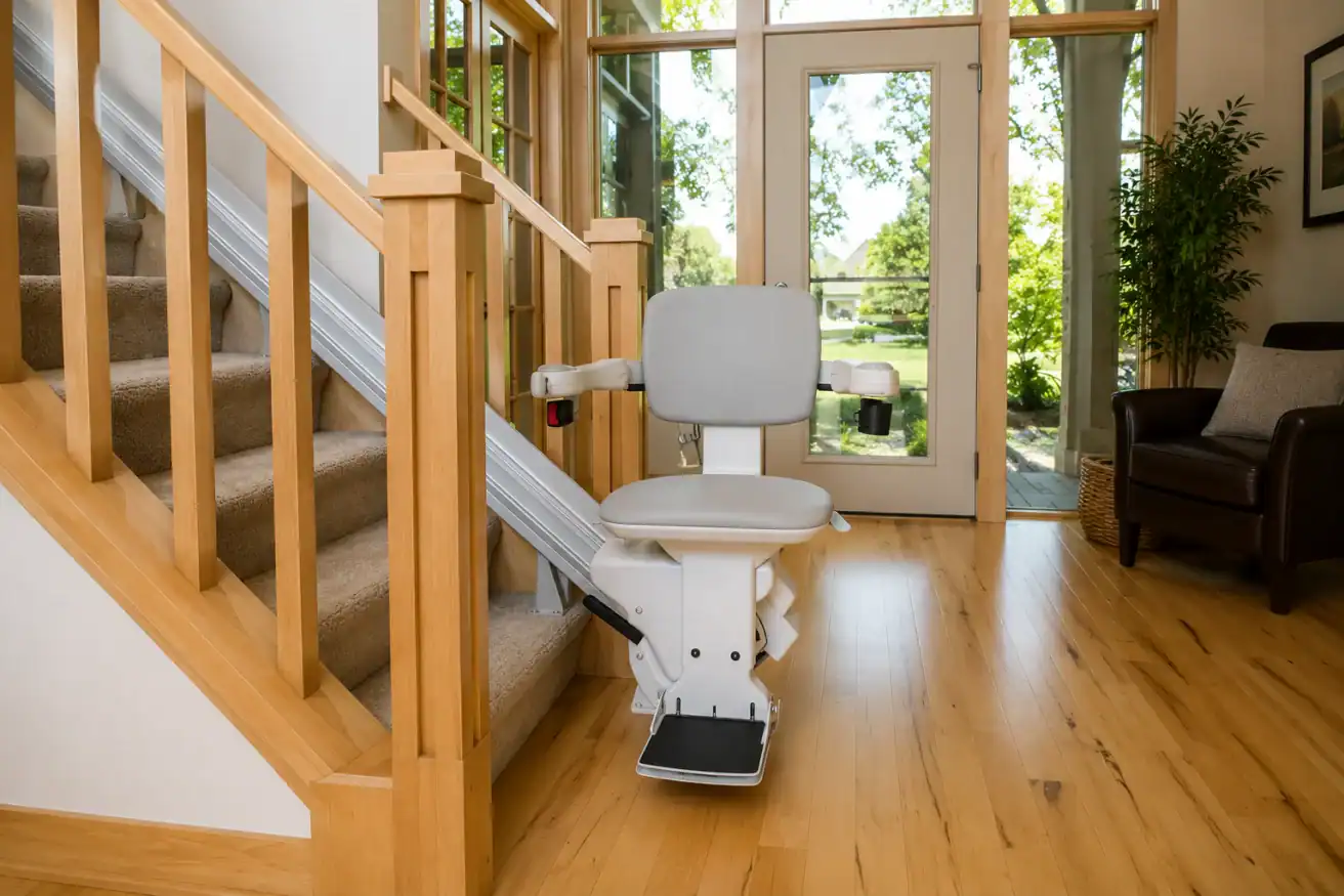 A stairlift is installed alongside a wooden staircase in a bright room with large windows, light wood floors, and a tan armchair.