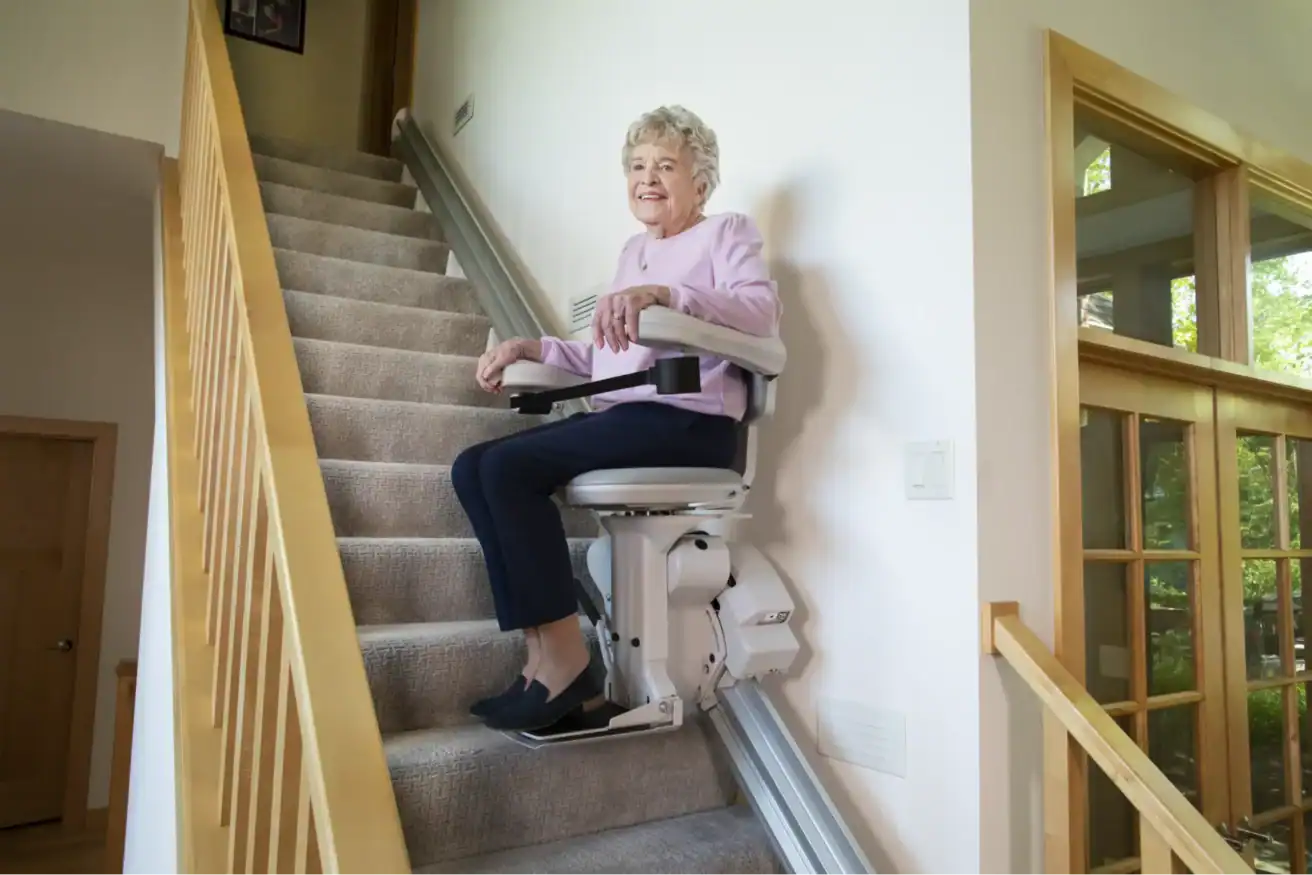 An older woman sits and rides up a staircase in a motorized stair lift chair inside a bright, modern home.