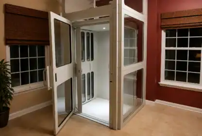 A residential elevator with open glass doors is shown in a room with tan tile flooring, red and beige walls, and large windows with brown blinds.