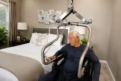 An older man in a wheelchair sits next to a bed under a ceiling lift, looking at a smartphone in a well-lit bedroom.