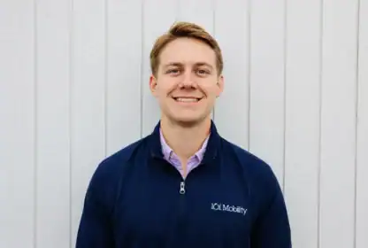 A man in a navy 101 Mobility jacket stands smiling in front of a light-colored wooden wall.