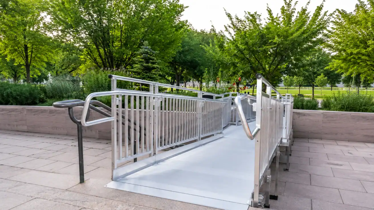 A metal accessibility ramp with railings is installed on a paved walkway surrounded by greenery and trees in an outdoor public area.