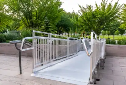 A metal accessibility ramp with railings is installed on a paved walkway surrounded by greenery and trees in an outdoor public area.